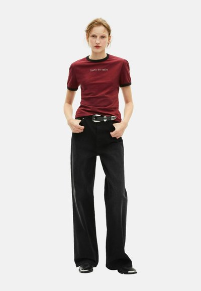 Rotes T-Shirt mit schwarzem Saum und Text, kombiniert mit hochgeschnittenen schwarzen weiten Hosen und glänzenden schwarzen Schuhen mit einem dekorativen Gürtel.