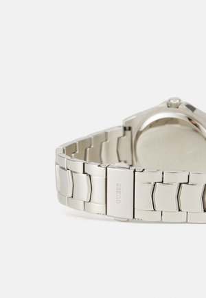 Srebrny zegarek metalowy z ogniwowym paskiem, posiadający polerowaną kopertę i minimalistyczny design. Zawiera zapięcie oznaczone "GUESS".