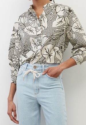 Frau trägt eine langärmelige Bluse mit schwarz-weißem Blumenmuster, die in eine hellblaue, hoch taillierte Jeans mit weißem Kordelgürtel gesteckt ist.