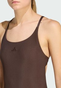 Mujer vestida con una camiseta de tirantes finos de color marrón ribeteada, que muestra el cuello, el hombro y la parte superior del brazo, contra un fondo claro y liso.