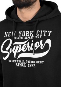 Crna dukserica s kapuljačom koja ima bijeli natpis: "NEW YORK CITY" i "Superior" u kurzivnom fontu, uz dizajn za košarkaški turnir, s vezicama.