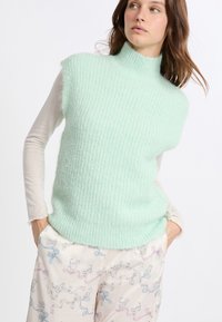 Maglione senza maniche di un verde menta con collo alto, caratterizzato da un design testurizzato e un materiale morbido e fluffy. Abbinato a pantaloni panna a fantasia.