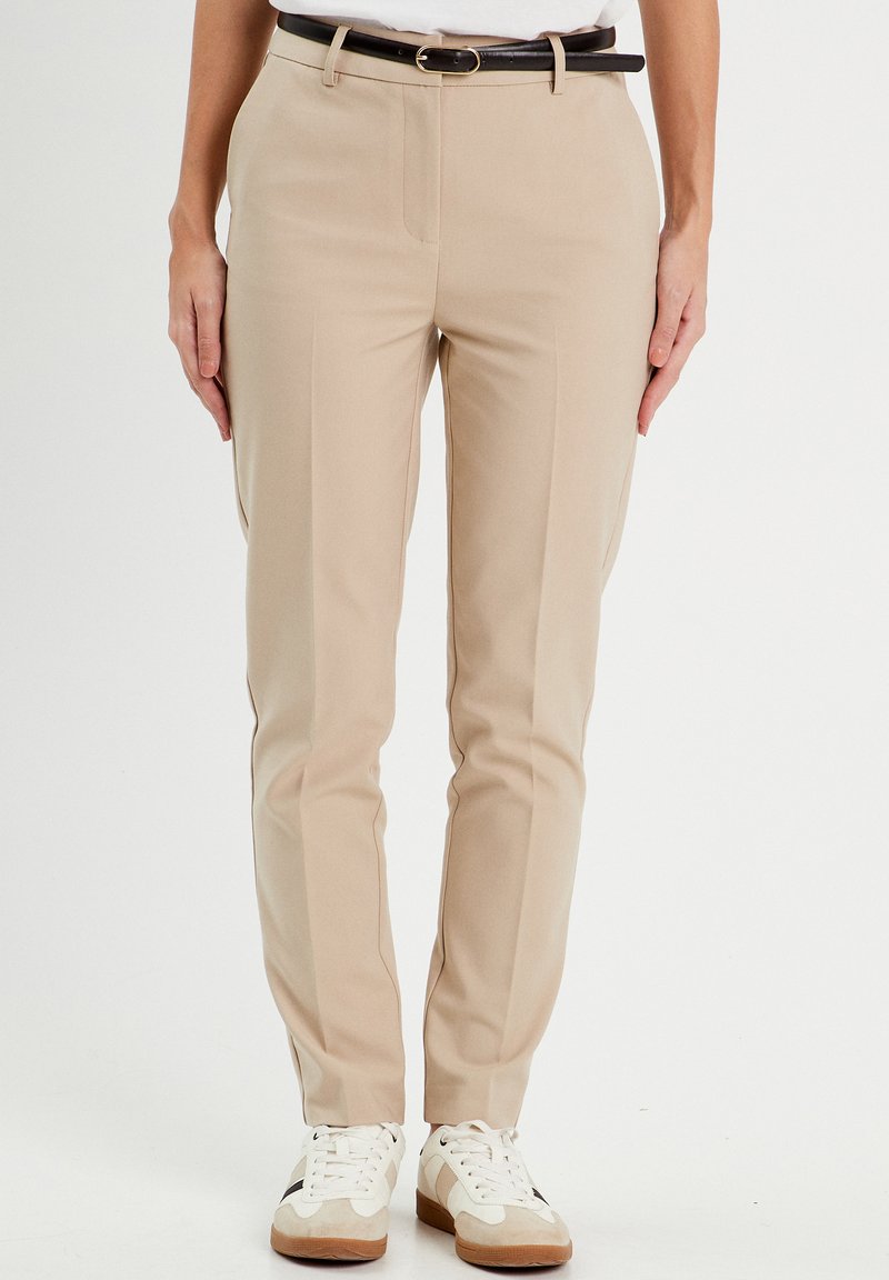 Beige tailored pants with a smooth texture, featuring front pockets and a black belt. Styled with white sneakers having tan accents.