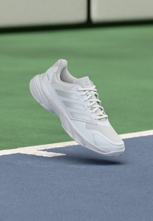 Baskets de sport blanches avec trois bandes et lacets, flottant au-dessus d'un court de tennis à surface verte et bleue.