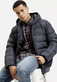 Granatowa puchowa kurtka z kapturem i przeszytymi panelami, noszona na koszuli w kratę. Całość uzupełniają casualowe niebieskie dżinsy i prosta krzesło.