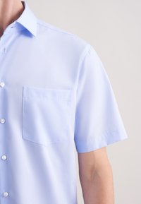 Jasnoniebieska koszula z krótkim rękawem wykonana z gładkiej tkaniny, z zapinanym na guziki przodem i płaską kieszenią na lewej stronie klatki piersiowej.