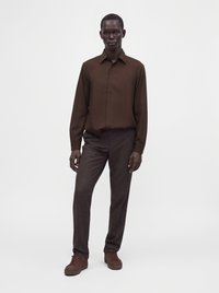 Modèle masculin debout, portant une chemise marron à manches longues, un pantalon marron foncé et des baskets marron sur un fond blanc uni.