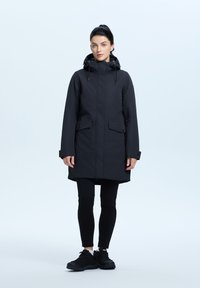 Parka nera impermeabile con colletto alto, cappuccio regolabile e due tasche frontali, abbinata a leggings neri e sneakers nere robuste.