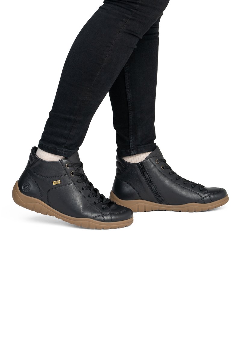 Personne portant des baskets montantes en cuir noir avec des semelles marron, un pantalon noir et des chaussettes blanches, debout avec un pied croisé devant l'autre.