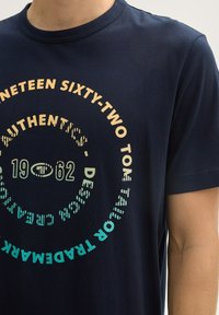 T-shirt en coton bleu marine avec un col rond, présentant un design graphique circulaire multicolore avec les inscriptions "Dix-neuf soixante-deux" et "Authentique".