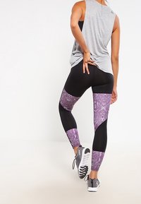 Legíny s černým základem a fialovými panely s hadím potiskem. Měkký, elastický materiál; doplněno volným šedým tílkem a sportovní obuví.