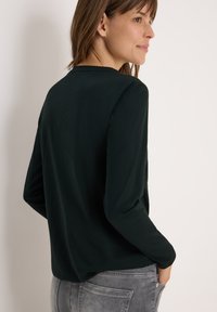 Haut à manches longues, de couleur vert foncé, en tissu doux. Présente un col rond et une coupe décontractée, associé à un jean gris clair dans un style décontracté.