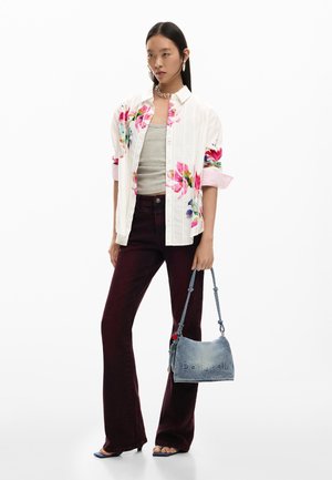 Femeie purtând o cămașă albă cu nasturi, cu model floral, peste un top gri, pantaloni evazați închiși la culoare, sandale albastre cu toc, ținând o geantă Desigual din denim.