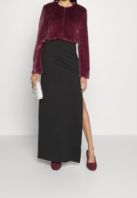 Veste courte en fausse fourrure bourgogne, robe maxi noire avec fente sur le côté, et chaussures bourgogne à enfiler texturées, accompagnées d'une pochette argentée.
