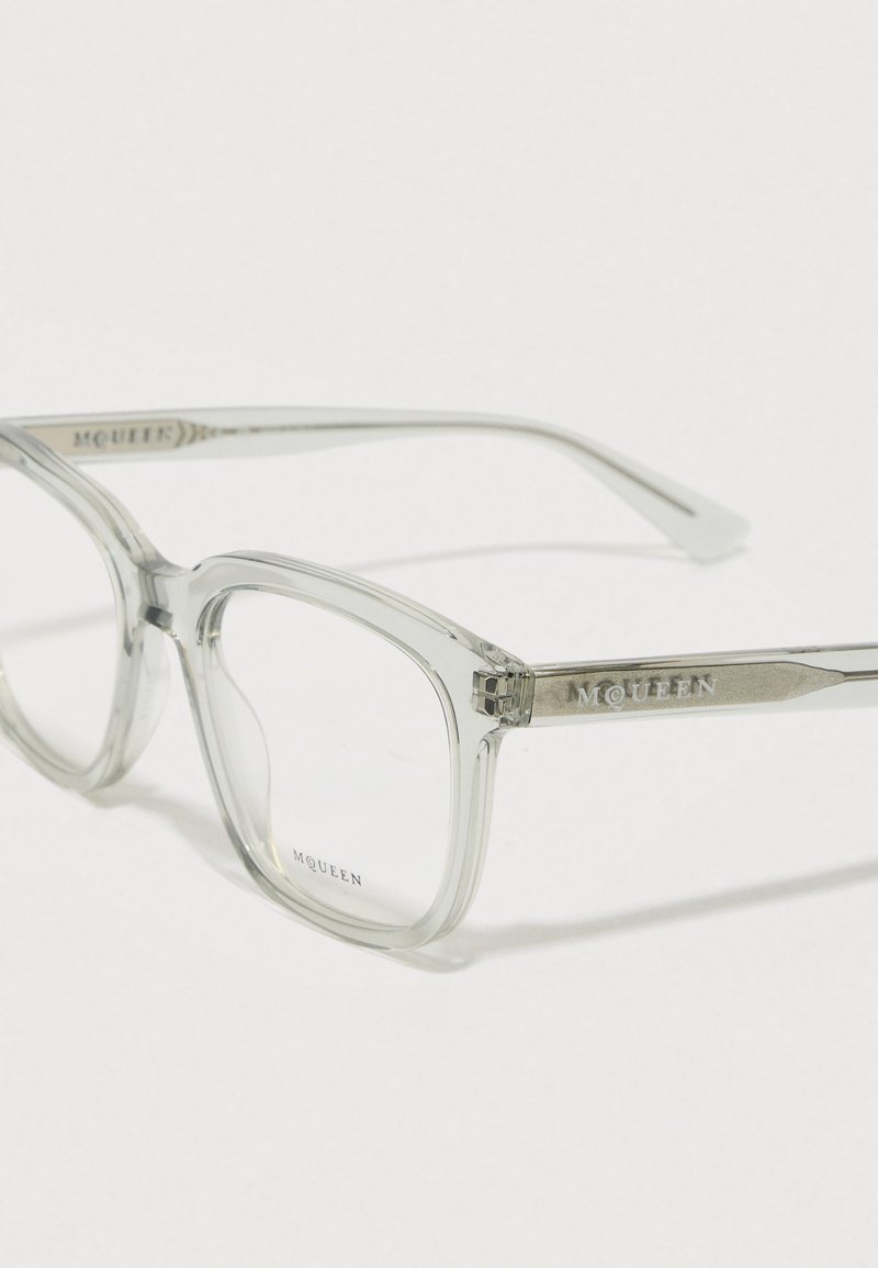 Gafas rectangulares transparentes con un marco claro. Patillas de metal con el logo "McQueen" grabado. Textura suave, lentes grandes.