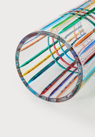 Verre en verre transparent avec des rayures verticales colorées en bleu, rouge, jaune, vert et orange, présenté incliné sur un fond blanc.