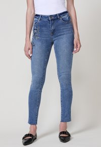 Jean en denim bleu skinny avec broderie florale sur la cuisse gauche, design à cinq poches, passants de ceinture et ourlet de cheville légèrement effiloché.