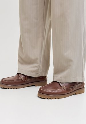 Scarpe in pelle marrone stile mocassino con suole di gomma color cognac indossate con pantaloni beige a righe sottili, in piedi su un pavimento bianco.