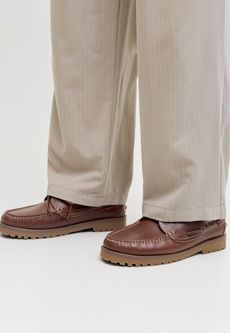 Chaussures mocassins en cuir marron avec semelles en caoutchouc beige portées avec un pantalon beige à rayures fines, debout sur un sol blanc.