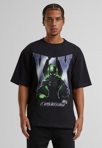 Musta ylipitkä t-paita, jossa on graafinen kuvasto vihreästä avaruusolennosta sekä tekstit "V1X" ja "OUTTA THIS WORLD" valkoisella ja sinisellä.