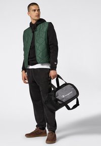 Gilet trapuntato verde sopra un hoodie nero, abbinato a pantaloni neri e sneakers marroni. Tiene una borsa a tracolla nera e grigia con il logo "Champion".