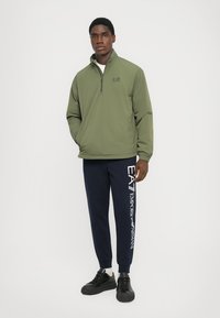 Oliven grønn halv-zip jakke i lett materiale; marineblå joggebukser med hvit EA7-logo; svarte treningssko med teksturerte såler.