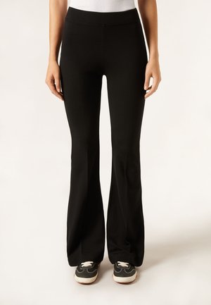 Kobieta w czarnych rozszerzanych spodniach i czarno-białych sportowych butach, stojąca na jednolitym tle.