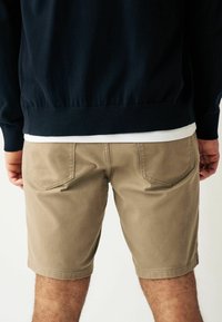 Shorts kaki avec une coupe classique, fabriqués en tissu mélangé coton. Dotés de poches arrière et de détails cousus. Portés avec un sweat-shirt bleu marine.