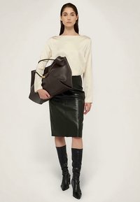 Schwarzer Lederminirock, cremefarbenes Langarmshirt und knielange schwarze Stiefel. Große dunkelbraune Handtasche mit goldenem Akzent in einer Hand gehalten.