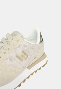 Scarpa sportiva beige con tomaia in rete, logo dorato e accento argento sul tallone. Caratterizzata da lacci rotondi e suola in gomma testurizzata.
