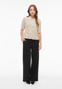 Beżowy dzianinowy kardigan z krótkim rękawem, z kołnierzykiem, zapinany na guziki oraz z dwiema przednimi kieszeniami. Połączony z czarnymi spodniami o wysokim stanie.