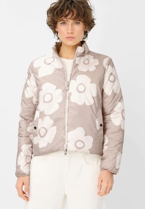 Frau trägt beige gesteppte Jacke mit großem weißem Blumenmuster, steht vor weißem einfarbigem Hintergrund.