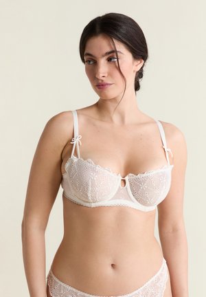 Kobieta ubrana w biały koronkowy biustonosz w kwiatowy wzór z fiszbinami i regulowanymi ramiączkami, patrząca w bok na jednolitym tle.