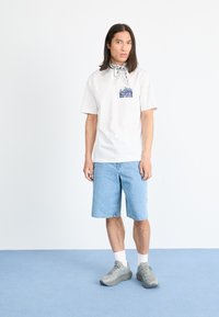 Hvit bomull T-skjorte med et blått grafisk trykk på brystet, kombinert med lyseblå denimshorts. Grå joggesko og et svart-hvitt mønstrede bandana.