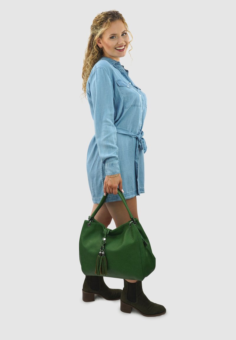Groene leren handtas met een textuurafwerking, voorzien van een kwastje en zilveren hardware, gedragen bij een blauwe denim shirtjurk en zwarte enkellaarsjes.