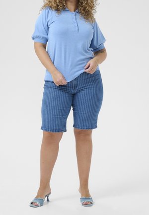 Kobieta o kręconych włosach, ubrana w niebieską koszulkę polo z krótkim rękawem, dżinsowe szorty w paski oraz jasnoniebieskie sandały na obcasie, stoi na białym tle.