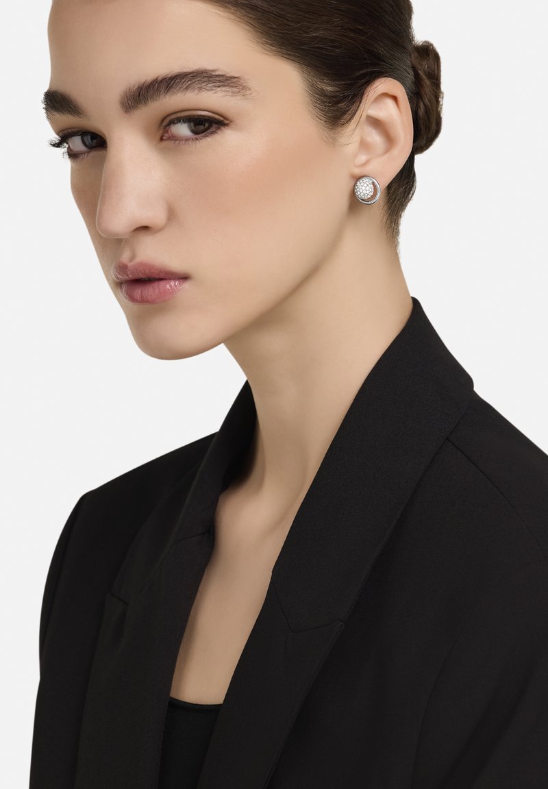 Sudraba auskari ar teksturētu virsmu, ko nēsā persona melnā žaketē, ar neitrālu fonu.