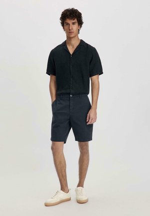 Chemise noire à manches courtes avec un col, associée à un short foncé. Portée avec des baskets crème, arborant un design simple et une texture lisse.