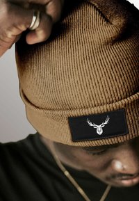 Brown ribbed beanie with a black rectangular patch featuring a white deer head logo. Close-up on fabric texture and fit on a head.