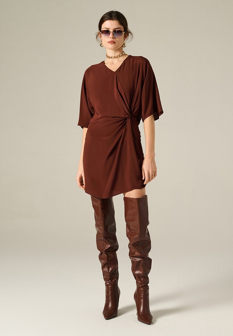 Abito corto marrone con tessuto drappeggiato e dettaglio annodato in vita. Abbinato a stivali alti marroni in pelle fin sopra il ginocchio e occhiali da sole.