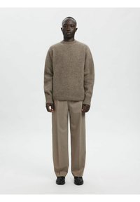 Brūns vilnas džemperis ar ribām aprocēs un apakšmalā, kombinēts ar gaiši bēšām plisētām biksēm un melnām kurpēm, radot atslābinātu siluetu.