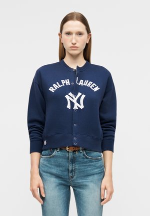 Kobieta o prostych, brązowych włosach, ubrana w granatową rozpinaną bluzę Ralph Lauren i jasnoniebieskie dżinsy, stoi na tle jednolitego tła.