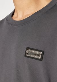 Grå bomull t-skjorte med rund hals, med en teksturert svart etikett med en sølvfarget Nike-logo på brystet. Glatt stofftekstur.