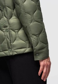 Uomo che indossa una giacca trapuntata verde oliva con bottoni a pressione sopra pantaloni neri, mostrando i dettagli delle maniche e del lato.