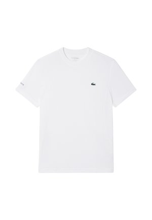 Camiseta blanca de manga corta con cuello redondo, logo pequeño de cocodrilo verde en el pecho izquierdo y texto "Lacoste" en la manga izquierda.