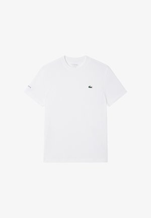 Camiseta blanca de manga corta con cuello redondo, logo pequeño de cocodrilo verde en el pecho izquierdo y texto "Lacoste" en la manga izquierda.