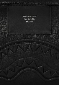 Bolso de cuero negro que presenta un diseño embosado de dientes de tiburón, superficie texturizada y una etiqueta de tela con "SPRAYGROUND Nueva York Est. 2010."