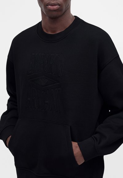 Άνδρας που φοράει μαύρο φούτερ Emporio Armani με ανάγλυφο λογότυπο και τσέπη καγκουρό, με τα χέρια μερικώς στις τσέπες.