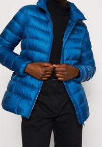 Blå puffjacka med en glansig yta, med horisontell sömnad, en hög krage och en framdragkedja. Bärs över en svart polotröja.