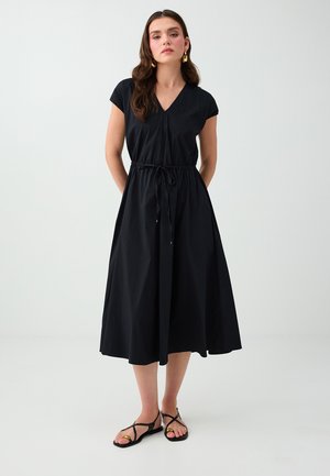 Femme aux longs cheveux bruns porte une robe midi noire avec une taille à cordon et des sandales plates noires à brides sur un fond blanc uni.